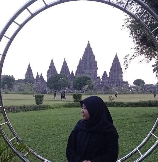 Gambar ini menunjukkan seorang wanita berhijab di depan candi yang megah. Dia memakai pakaian hitam dan berdiri di tengah taman. Dia tersenyum dan melihat ke arah candi. Terdapat kerangka besi setengah lingkaran di sekitar wanita itu. Di bagian belakang wanita tersebut, terdapat candi yang megah dengan arsitektur yang rumit. Candi tersebut dikelilingi oleh taman hijau yang luas. Di bagian depan candi terdapat beberapa orang yang sedang berjalan di taman. Atmosfer di gambar ini menenangkan dan damai.