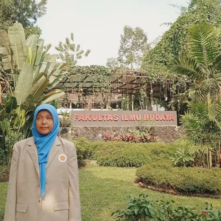 Gambar ini menunjukkan seorang wanita berhijab berdiri di depan sebuah bangunan dengan tulisan "FAKULTAS ILMU BUDAYA". Wanita itu mengenakan blazer berwarna coklat muda dan kerudung biru. Ia tampak tersenyum ke arah kamera. Di belakangnya terlihat taman dengan tanaman hijau yang rimbun. Bangunan tersebut memiliki arsitektur yang unik dengan dinding batu dan atap dari kayu.